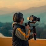 A woman holding a camera with gimbal captures the serene mountain scenery at dusk.