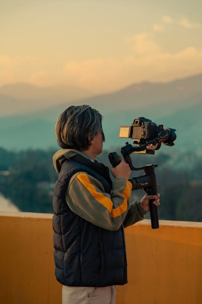 A woman holding a camera with gimbal captures the serene mountain scenery at dusk.